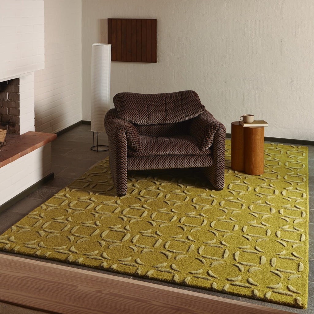A mustard yellow and beige patterned area rug in a mid-century modern living room setting, styled with a brown armchair, a wooden side table, and a fireplace.