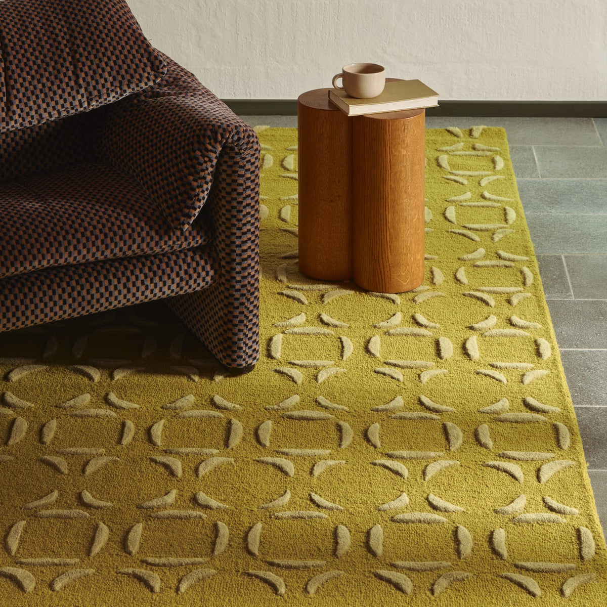 A mustard yellow area rug with a raised cream geometric pattern, styled in a living room setting with a brown patterned armchair and a wooden side table.