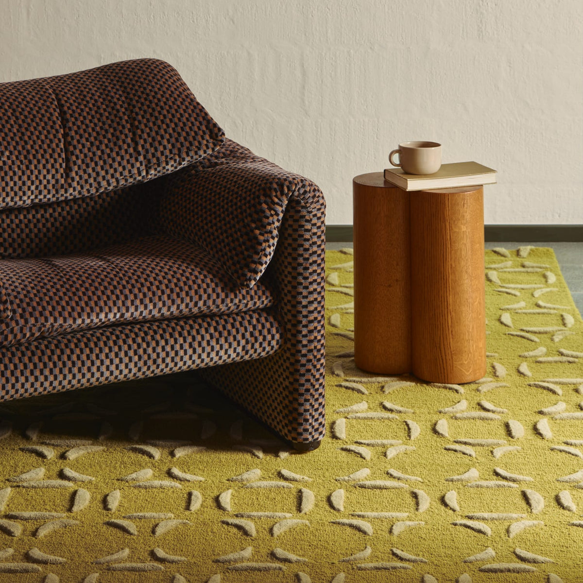 A modern brown patterned armchair sits beside a two-bench wooden side table holding a small cup, all placed on a textured yellow rug against a plain off-white wall.