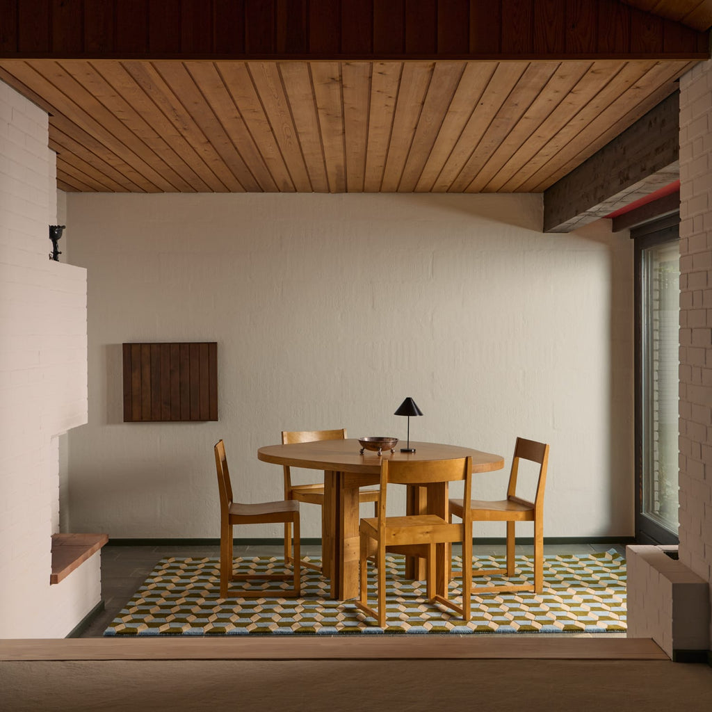 A mid-century modern dining set consisting of a round wooden table and four matching wooden chairs, styled on a geometric patterned rug in a minimalist room with a wood-paneled ceiling.