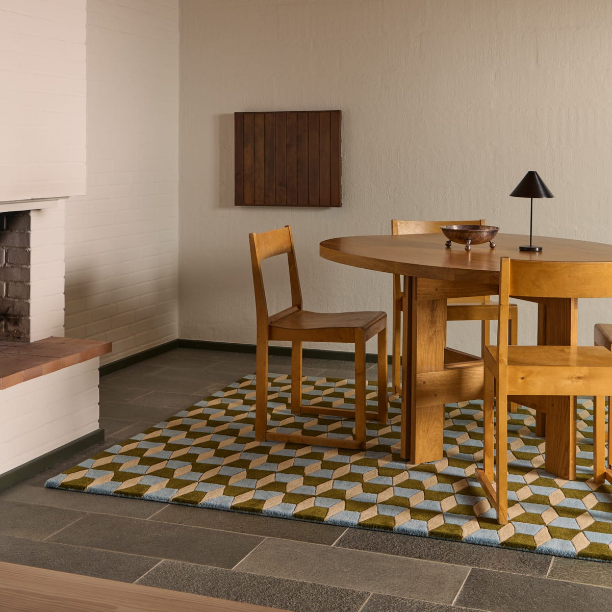 A modern dining room scene featuring a round wooden table and four matching wooden chairs placed on a geometric patterned area rug. The room has textured off-white walls, a dark tiled floor, and a white brick fireplace on the left.