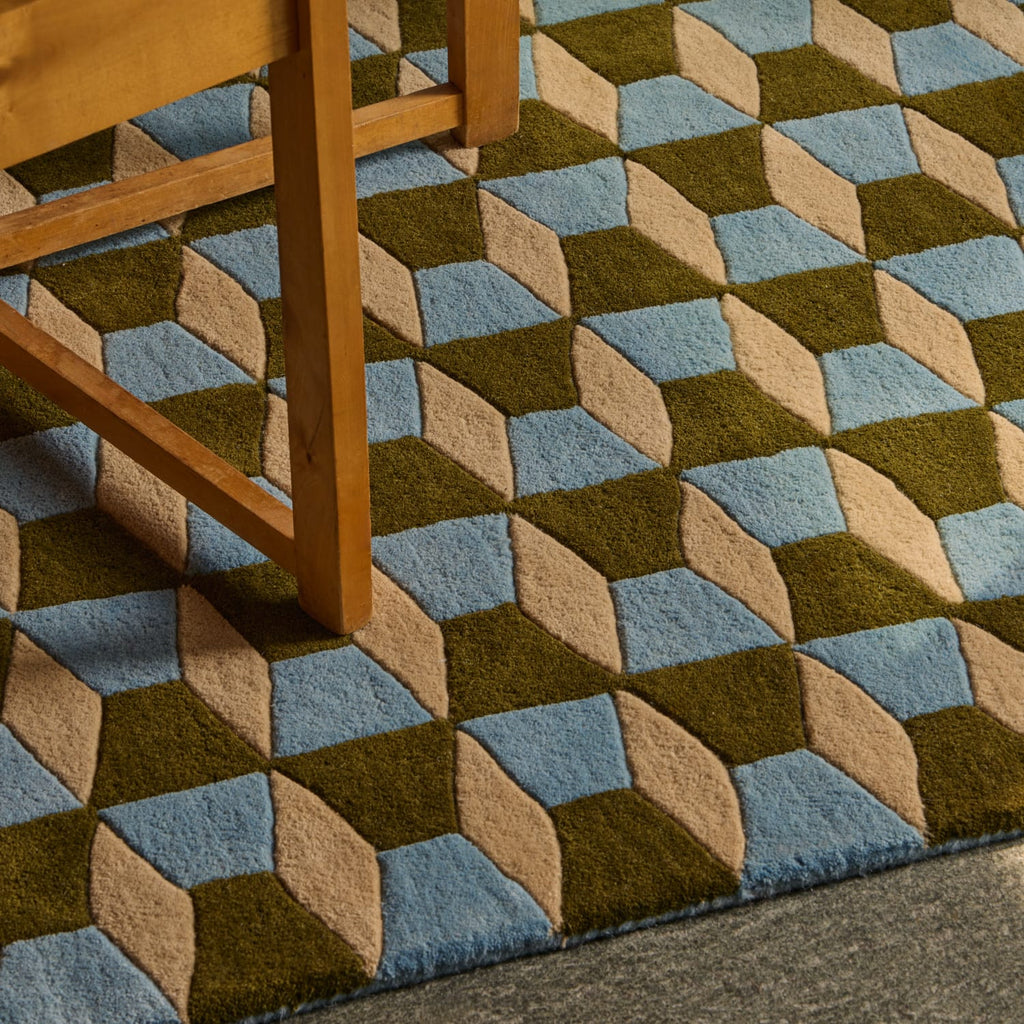 A close-up of a textured area rug with a 3D geometric cube pattern in olive green, light blue, and beige, shown under the legs of a wooden chair.