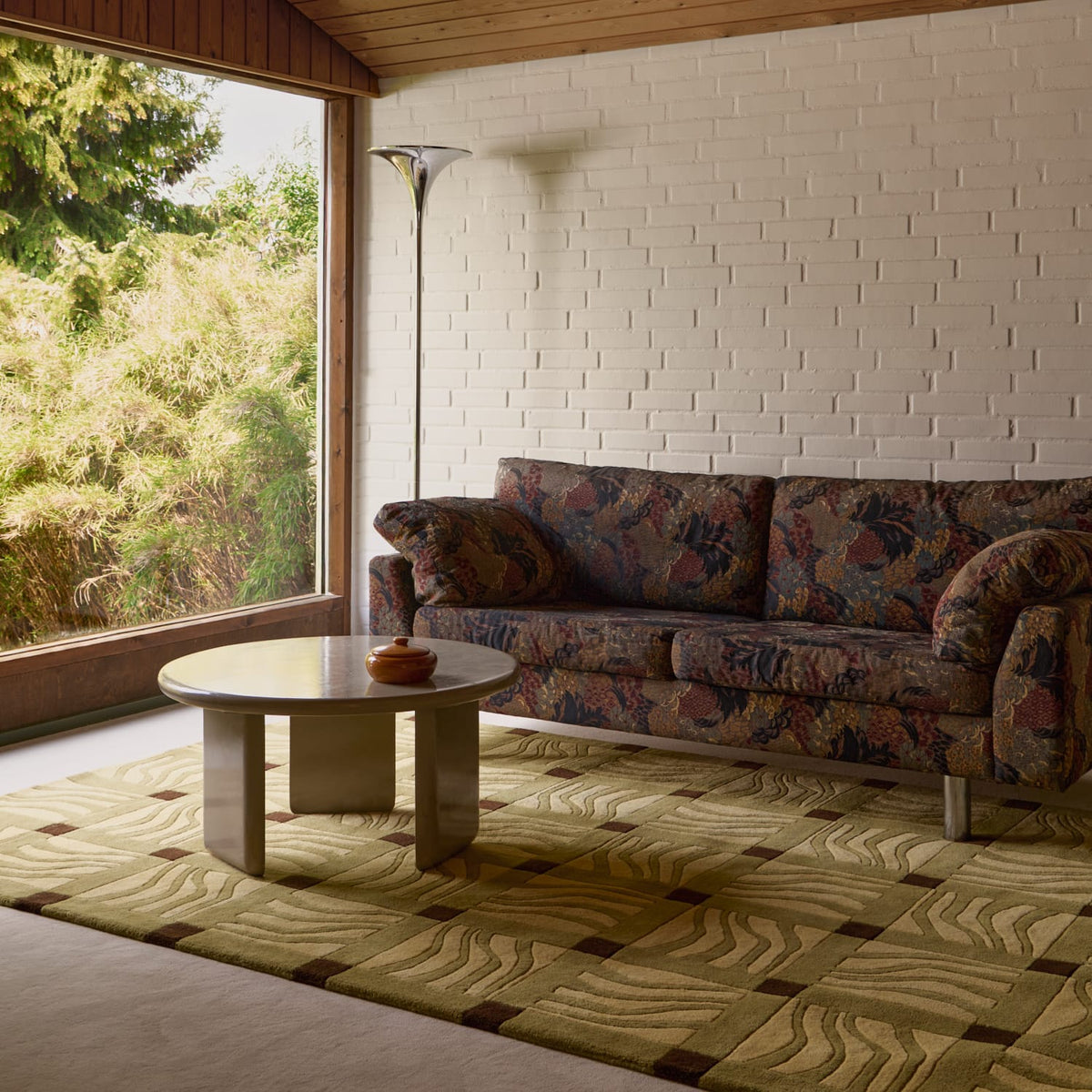A mid-century modern living room featuring a floral patterned three-seater sofa, a round glass-top coffee table, a geometric patterned rug, and a tall silver floor lamp against a white brick wall.