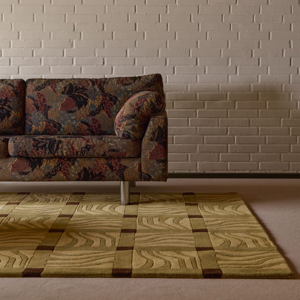 A close-up of a textured area rug with a wavy wood-grain pattern in beige, olive green, and brown, styled in a living room setting under a floral-patterned sofa against a white brick wall.