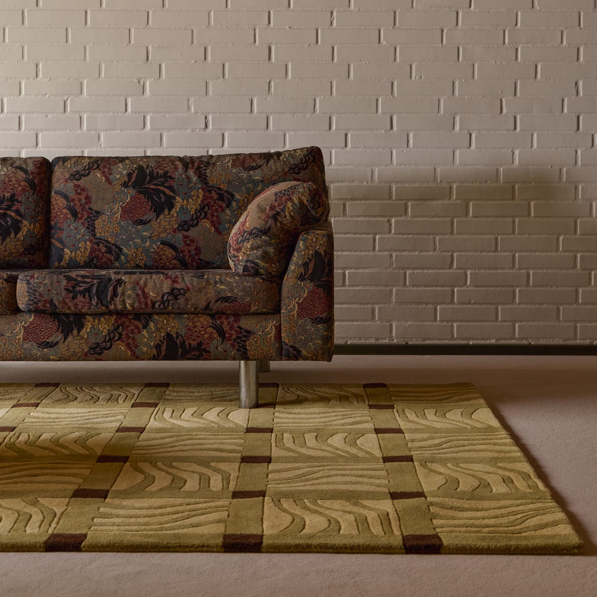 A close-up of a textured area rug with a wavy wood-grain pattern in beige, olive green, and brown, styled in a living room setting under a floral-patterned sofa against a white brick wall.