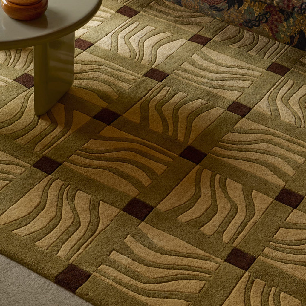 A close-up of a patterned area rug with a geometric grid design, featuring wavy stripe motifs in beige and olive green with dark brown square accents, shown in a styled interior setting with a table leg visible.