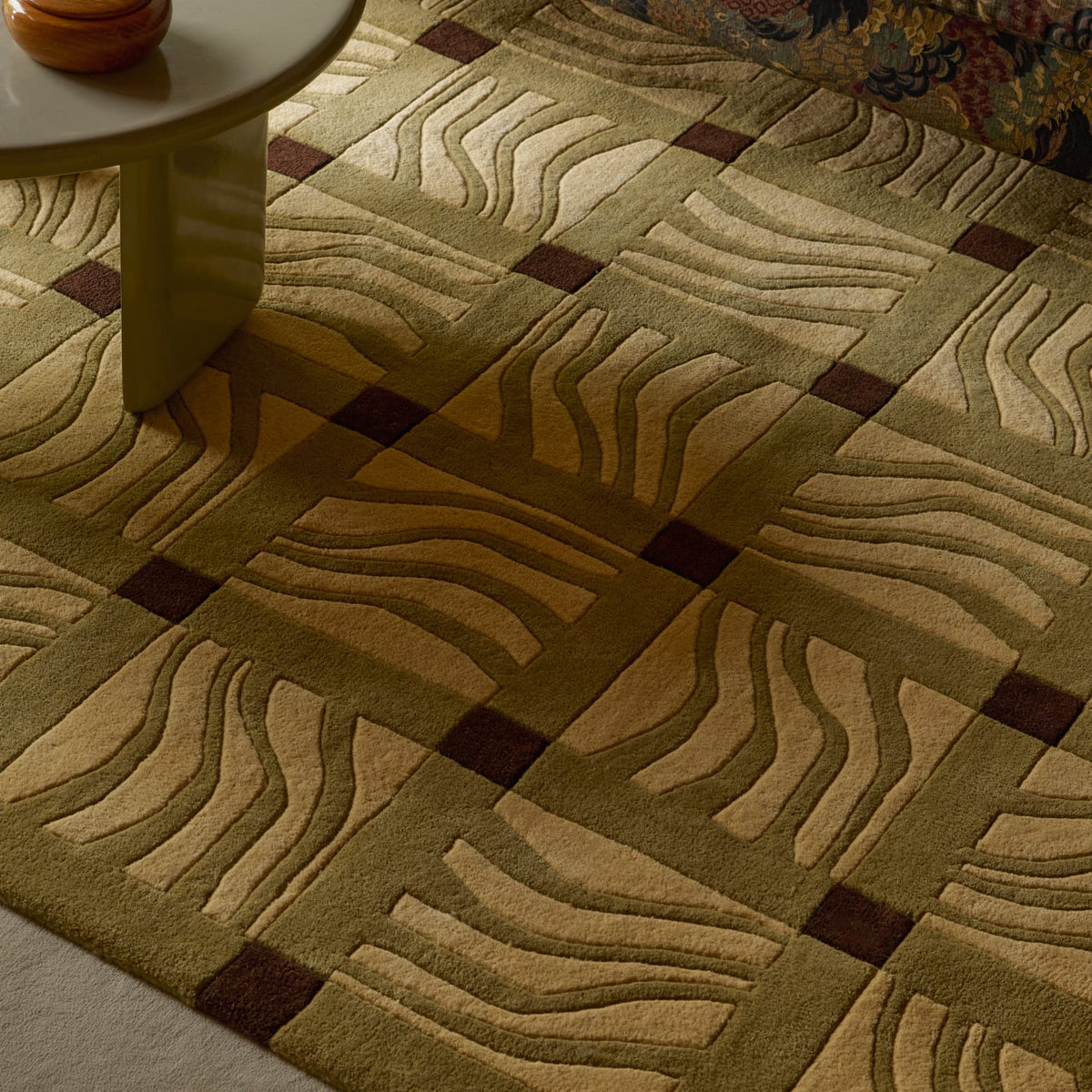 A close-up of a patterned area rug with a geometric grid design, featuring wavy stripe motifs in beige and olive green with dark brown square accents, shown in a styled interior setting with a table leg visible.