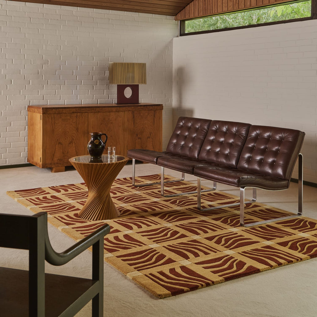 A mid-century modern living room scene featuring a dark brown tufted leather three-seater sofa on a patterned brown and beige rug, with a wooden sideboard, table lamp, and wood-paneled ceiling in the background.