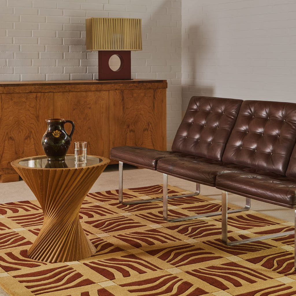 A mid-century modern living room scene featuring a brown tufted leather sofa, a sculptural fluted wood side table, a patterned brown and beige rug, and a wooden credenza against a white brick wall.