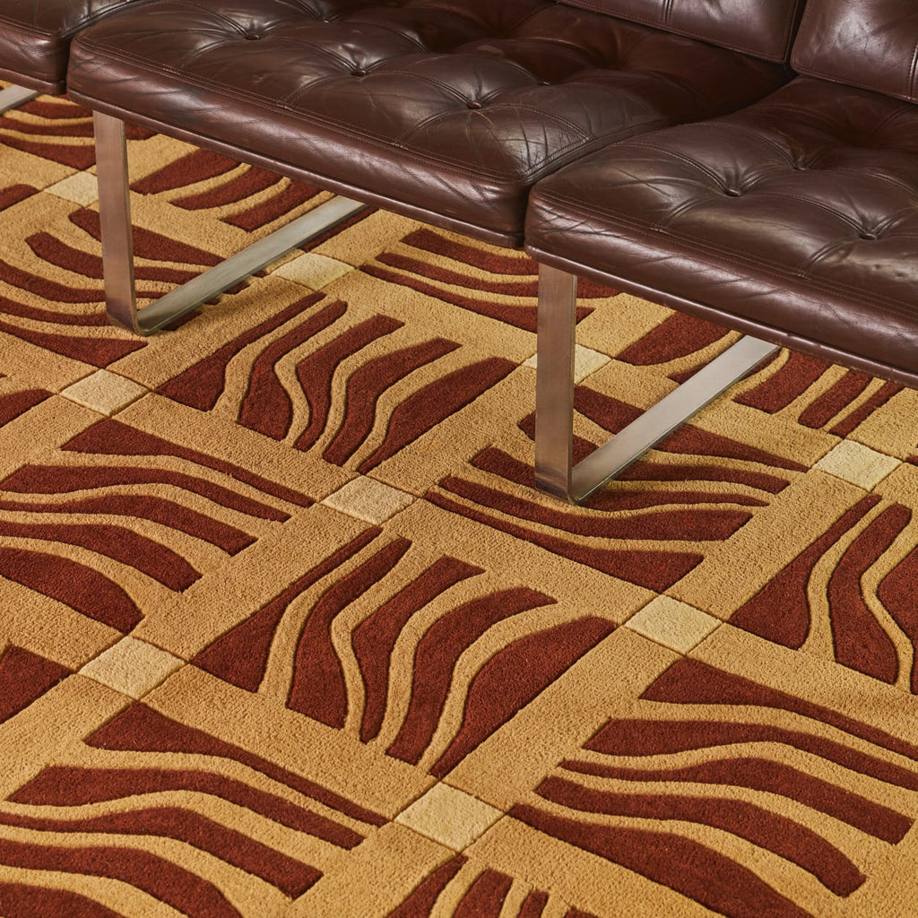 A close-up of a patterned area rug with a geometric wavy design in brown and beige, shown in a modern room setting with a dark brown leather sofa.