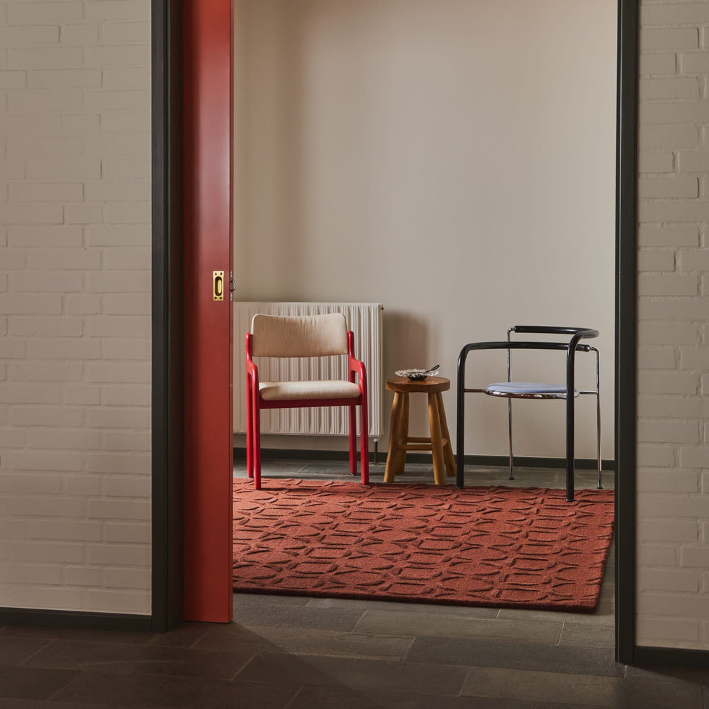 A red textured area rug with a geometric pattern, styled in a minimalist room with two chairs, a wooden stool, and a red door frame visible in the foreground.