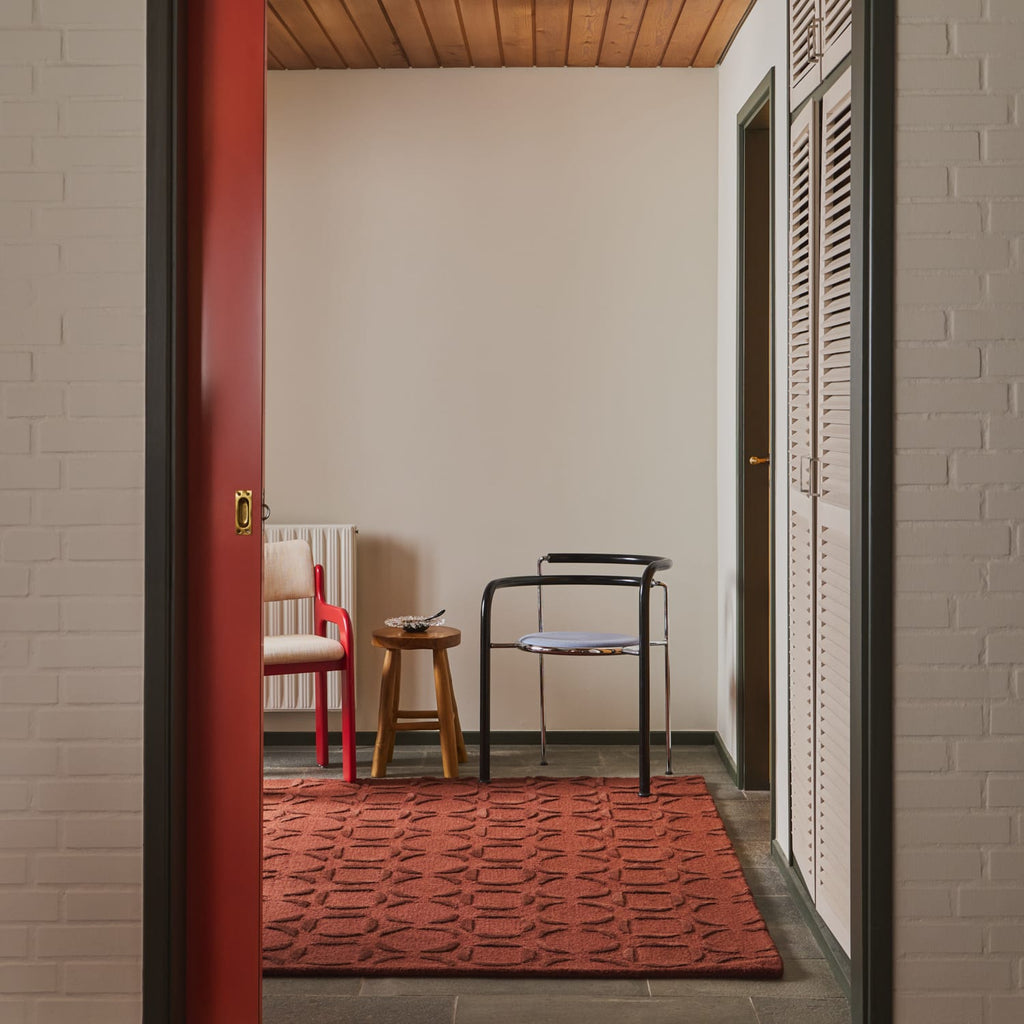 A rust-red-colored textured area rug laid out on a gray tiled floor in a modern interior hallway, framed by a red door on the left and white brick walls on the right.