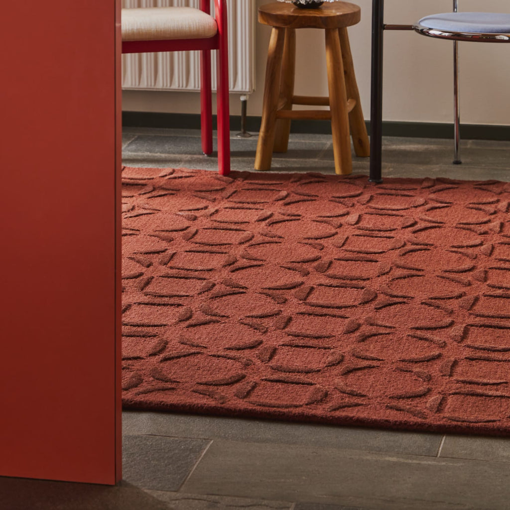 A rust-red textured area rug with a repeating geometric pattern, shown on a dark tiled floor in a room setting with furniture partially visible.