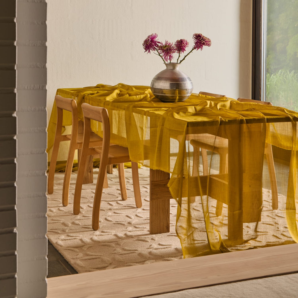 A bright yellow sheer tablecloth draped over a wooden dining table with matching wooden chairs, styled with a metallic vase of pink flowers on a light rug in a modern interior setting.
