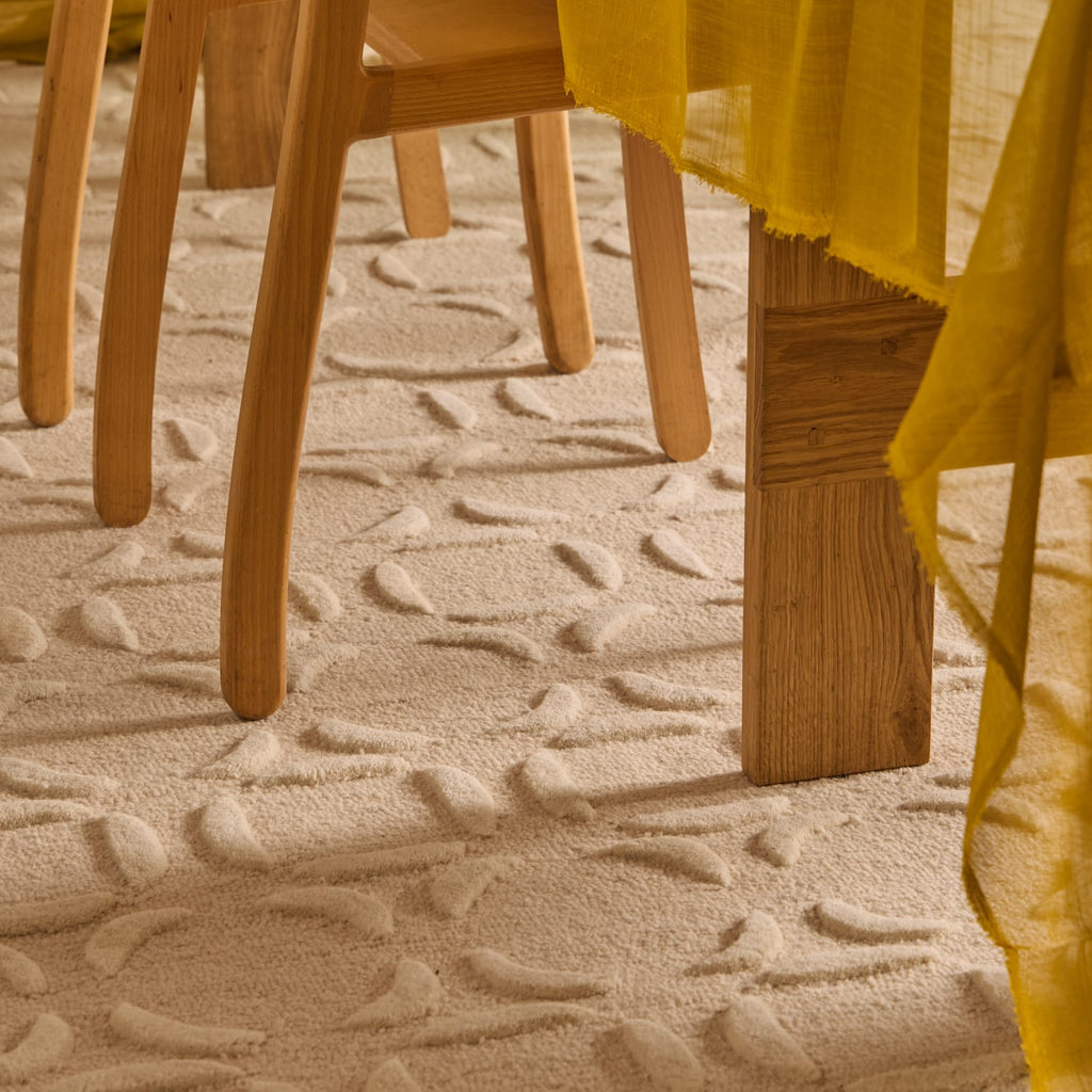 Close-up of a textured cream-colored area rug with a raised leaf pattern, shown under the legs of light wood dining chairs with a yellow tablecloth draped over the side.