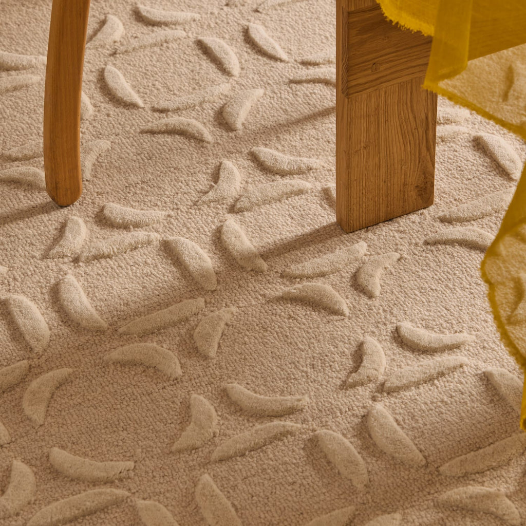 Close-up of a textured beige area rug with a raised leaf pattern, shown under the legs of wooden furniture with a yellow fabric drape visible on the right.