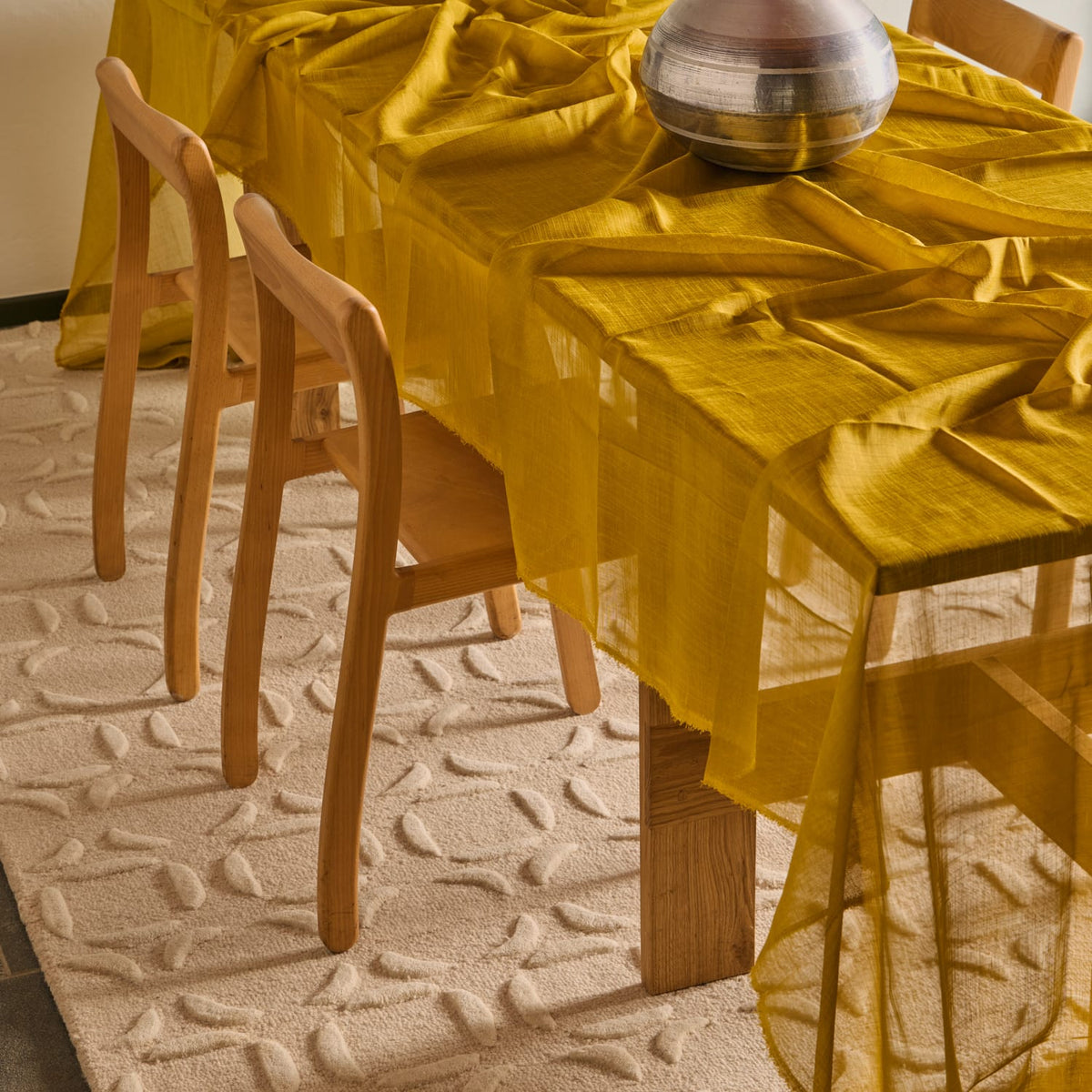 A sheer mustard yellow tablecloth draped over a wooden dining table, with matching chairs and a textured cream rug underneath.