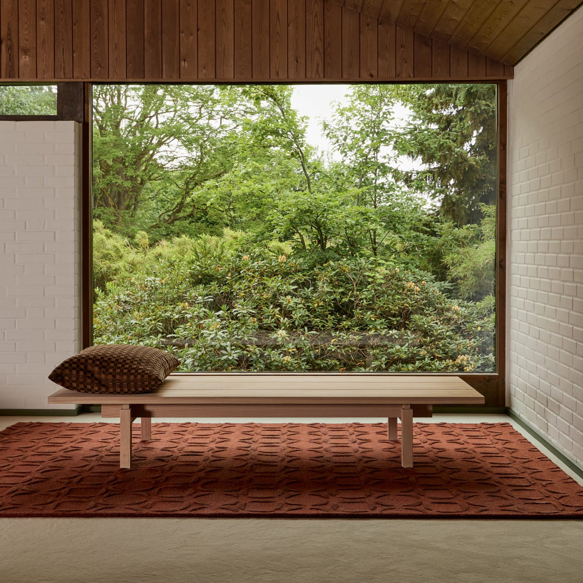 A low light-wood bench with a brown patterned throw pillow sits on a rust-red textured rug in a modern room with white brick walls, wood ceiling panels, and a large window overlooking a lush green garden.
