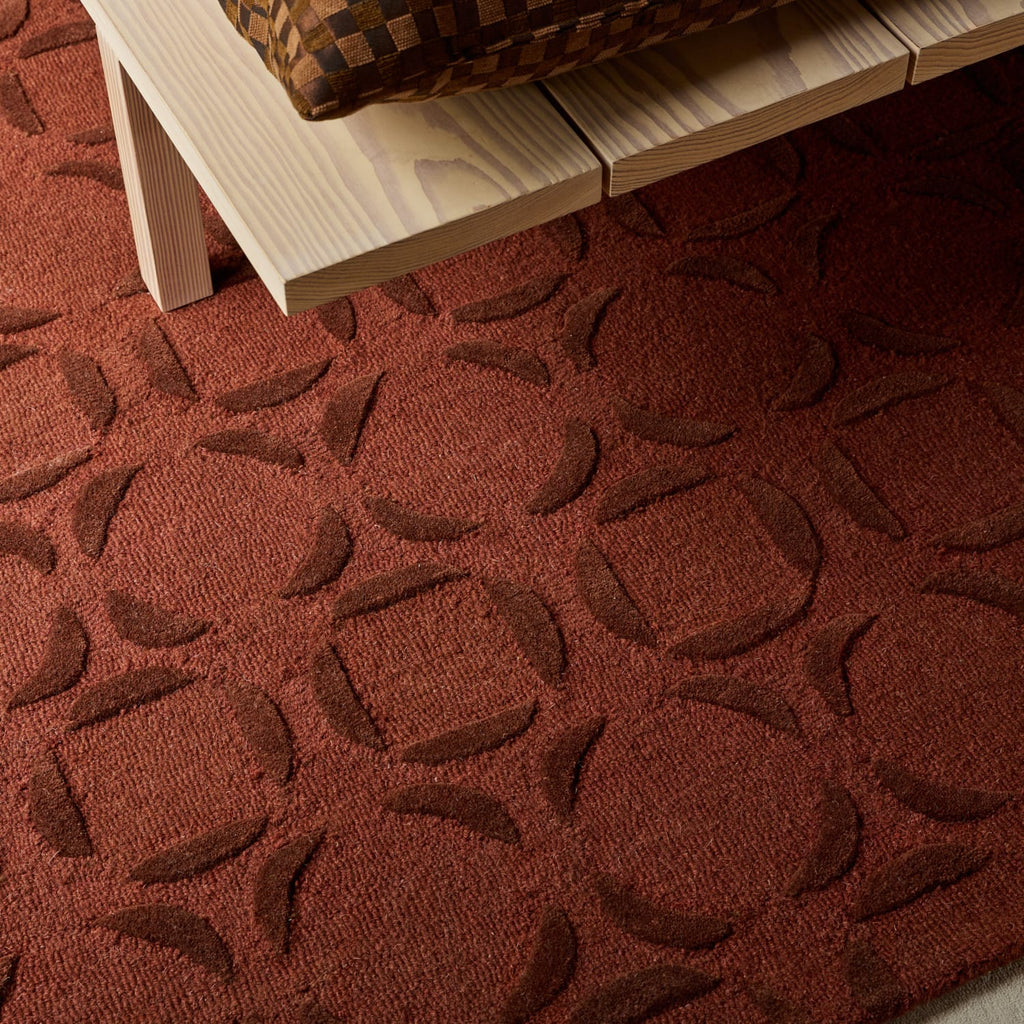 A close-up of a rust red area rug with a raised geometric pattern, shown under a light wood bench with a woven cushion.