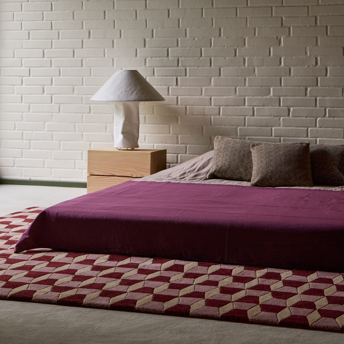 A bed dressed in a deep plum-purple comforter with three textured brown pillows, set in a bedroom with a white brick wall, a light wood nightstand, and a geometric patterned rug.