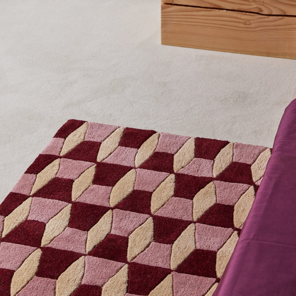 A close-up of a textured area rug with a 3D geometric cube pattern in burgundy, dusty pink, and beige, styled on a light-colored carpet next to a purple upholstered chair.