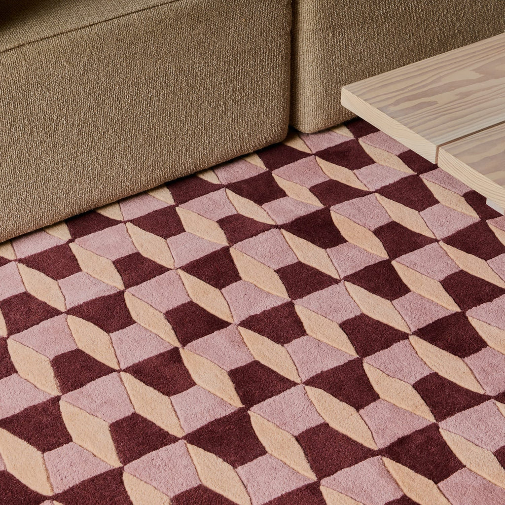 A geometric area rug with a 3D cube pattern in dusty pink, burgundy, and beige, styled in a living room setting on the floor between a brown sofa and a light wood table.