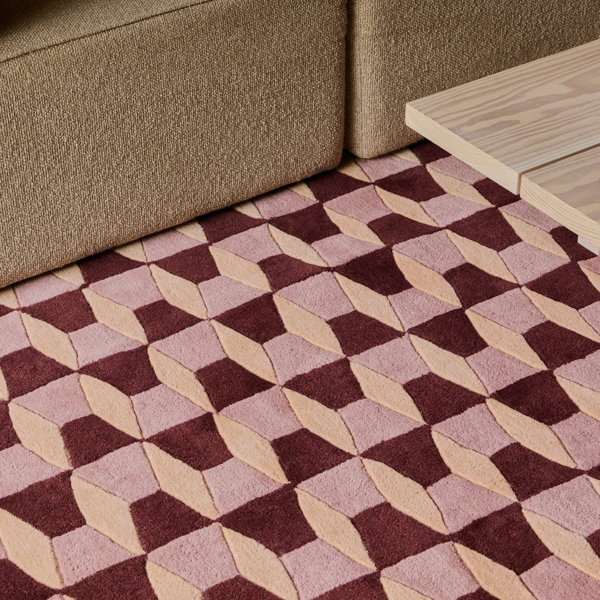 A geometric area rug with a 3D cube pattern in dusty pink, burgundy, and beige, styled in a living room setting on the floor between a brown sofa and a light wood table.