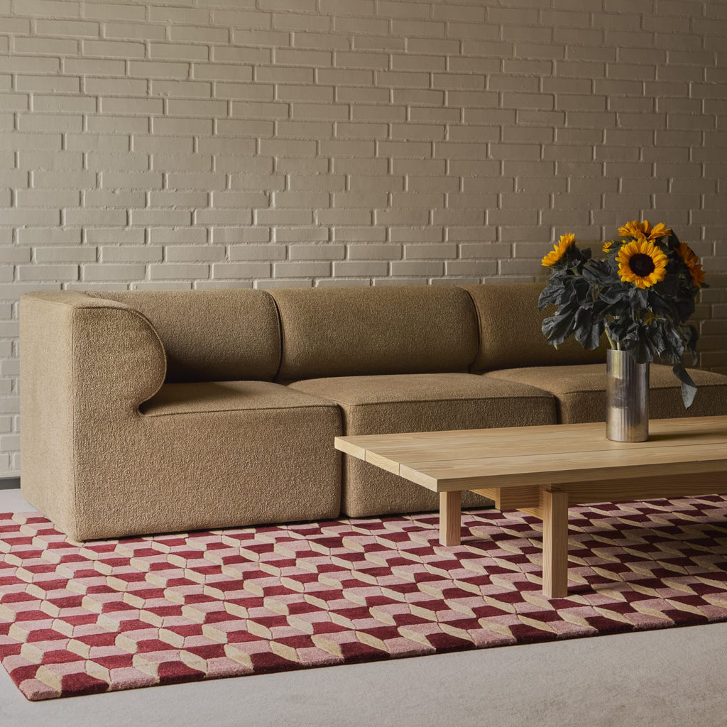 A modern beige upholstered sofa set against a light brick wall, with a light wood coffee table and a red and beige geometric patterned rug in the foreground. A vase of sunflowers sits on the table.