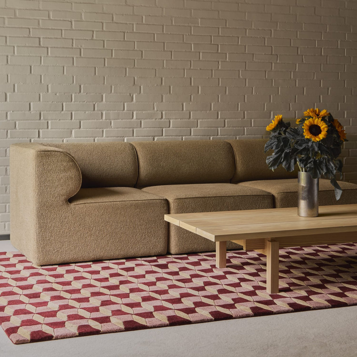 A modern beige upholstered sofa set against a light brick wall, with a light wood coffee table and a red and beige geometric patterned rug in the foreground. A vase of sunflowers sits on the table.