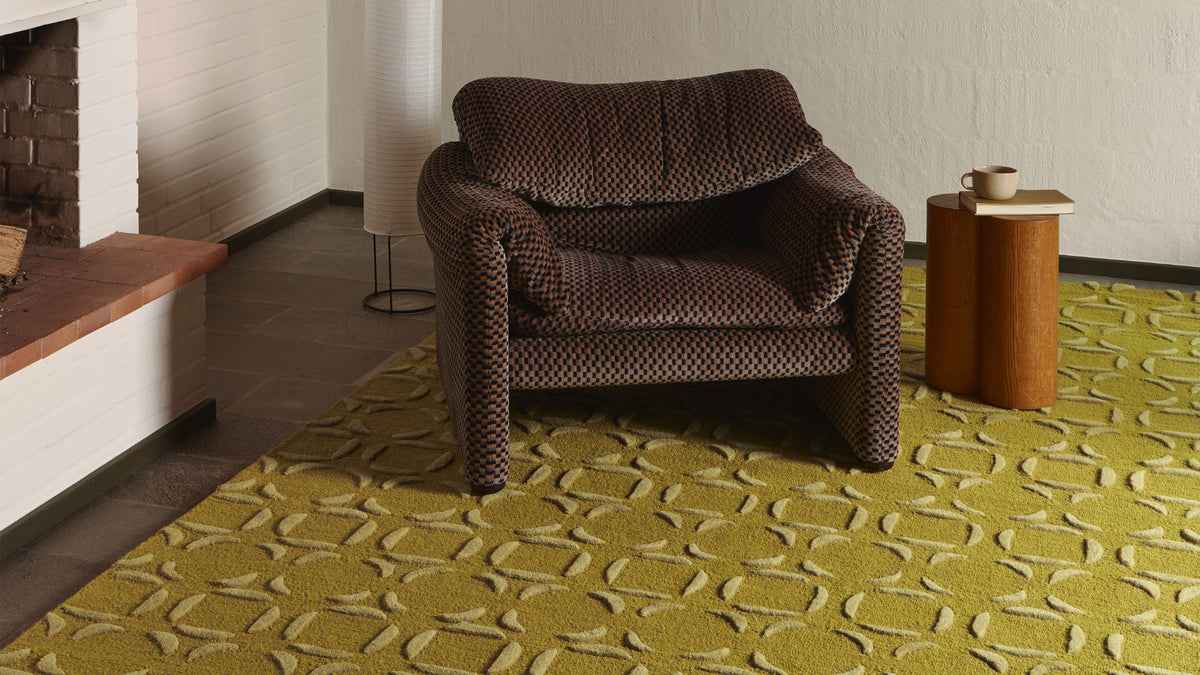 A mid-century modern armchair upholstered in a brown and black geometric patterned fabric, set on a yellow-green patterned area rug in a styled living room.