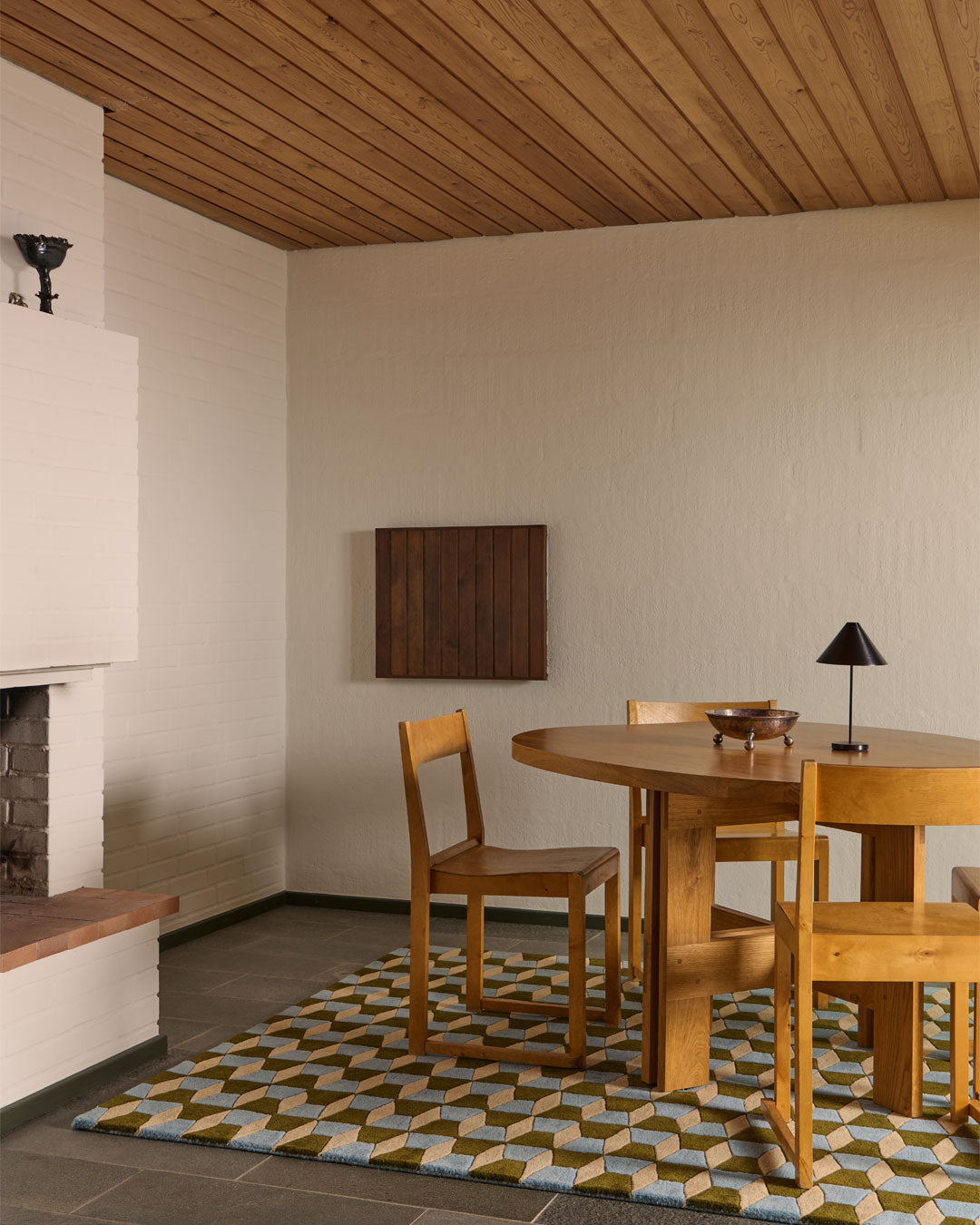 A mid-century modern dining room corner featuring a round wooden dining table and four matching wooden chairs on a geometric patterned rug. The room has textured off-white walls, a wood-paneled ceiling, and a white fireplace on the left.