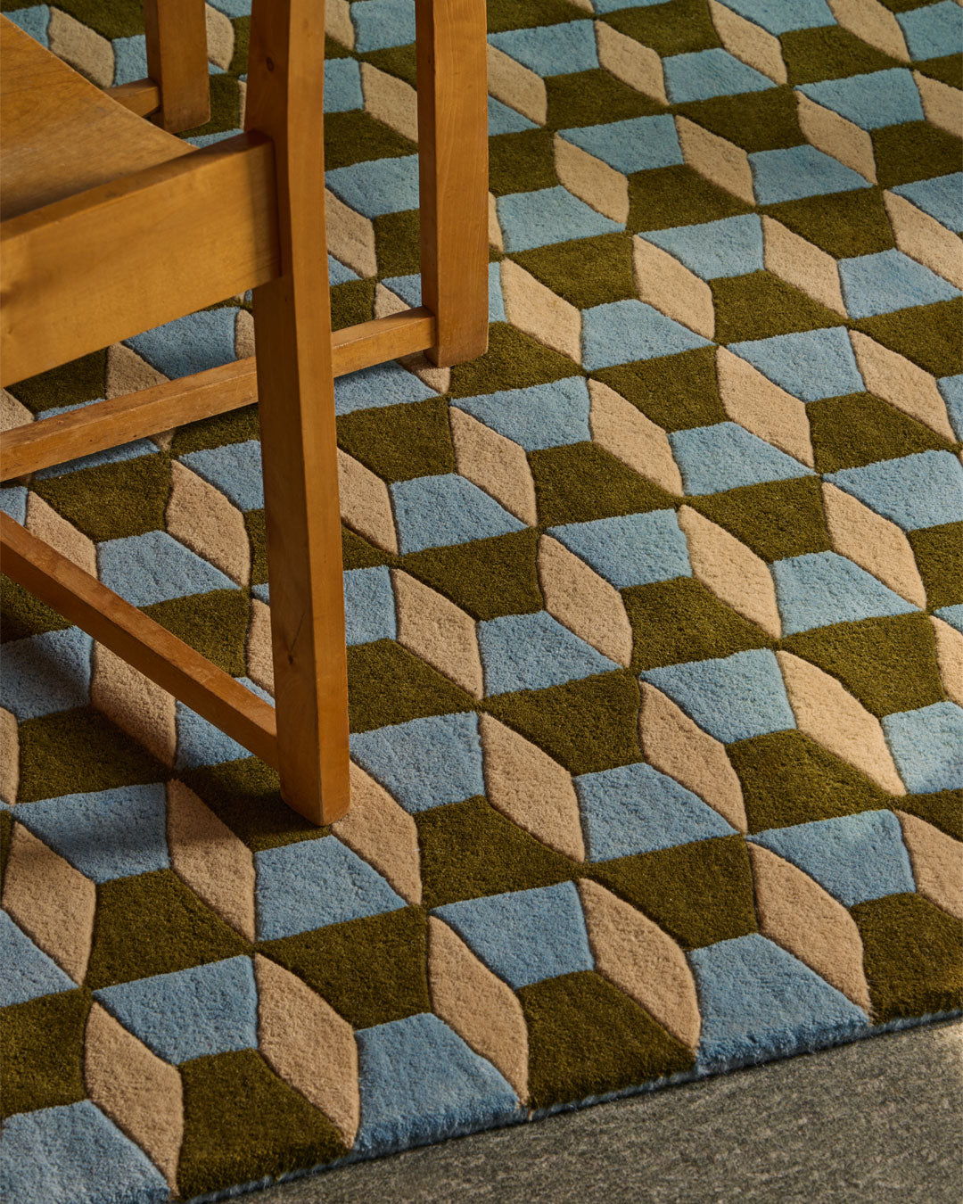 A close-up of a textured area rug with a 3D geometric cube pattern in olive green, light blue, and beige, shown with the legs of a wooden chair resting on it.