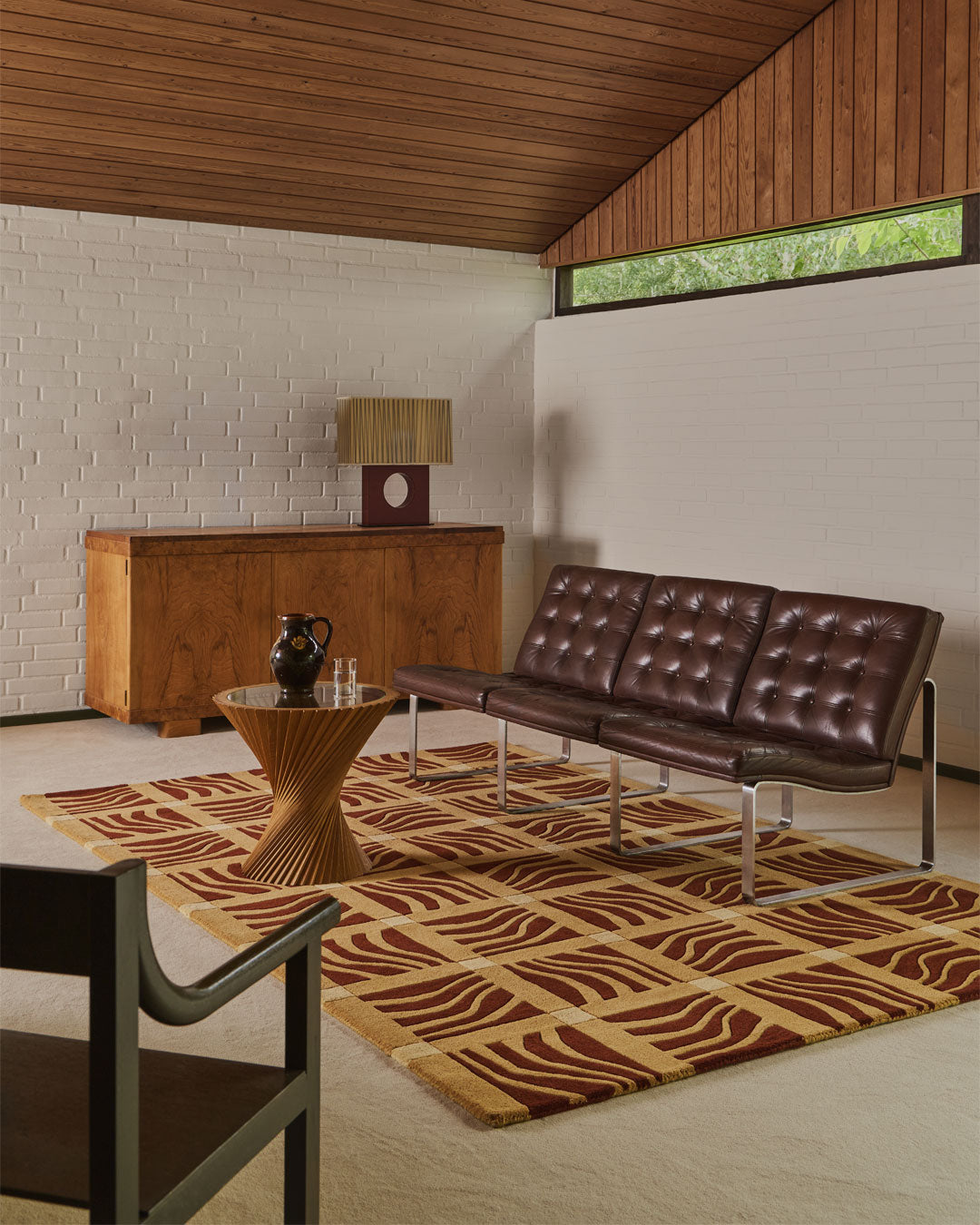A mid-century modern living room featuring a brown tufted leather sofa on a patterned brown and beige rug, with a wooden sideboard, wood ceiling, and white brick wall.