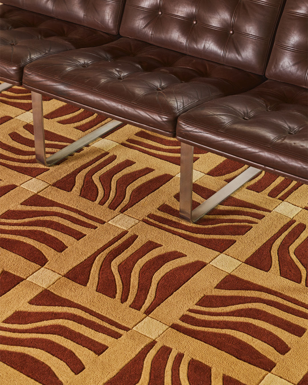 A close-up of a brown tufted leather sofa with metal legs placed on a patterned area rug featuring wavy stripes in brown and tan.