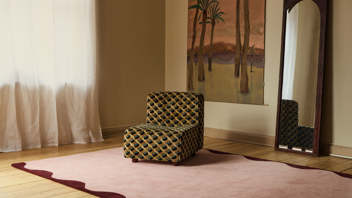A small upholstered armchair with a black and yellow geometric pattern sits on a pink rug in a styled room with wood floors, a mirror, and a painting.