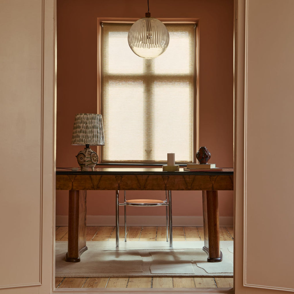 A warm, rustic wooden writing desk in a beautifully styled room with dusty rose walls, a window with a beige roller shade, and a light-colored rug.