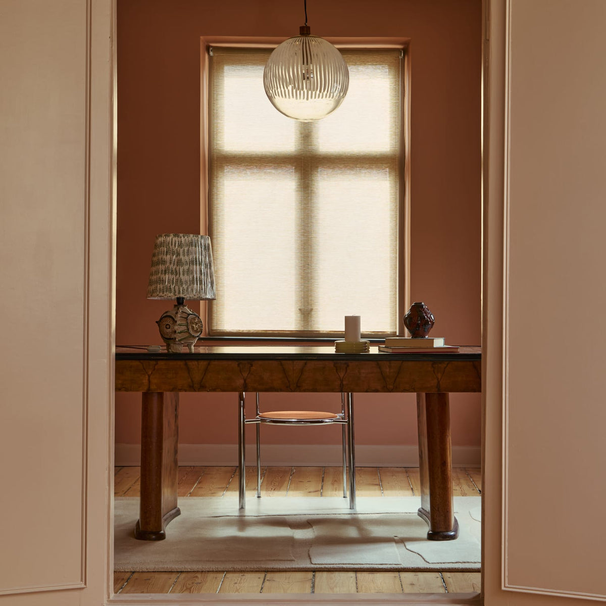 A warm, rustic wooden writing desk in a beautifully styled room with dusty rose walls, a window with a beige roller shade, and a light-colored rug.