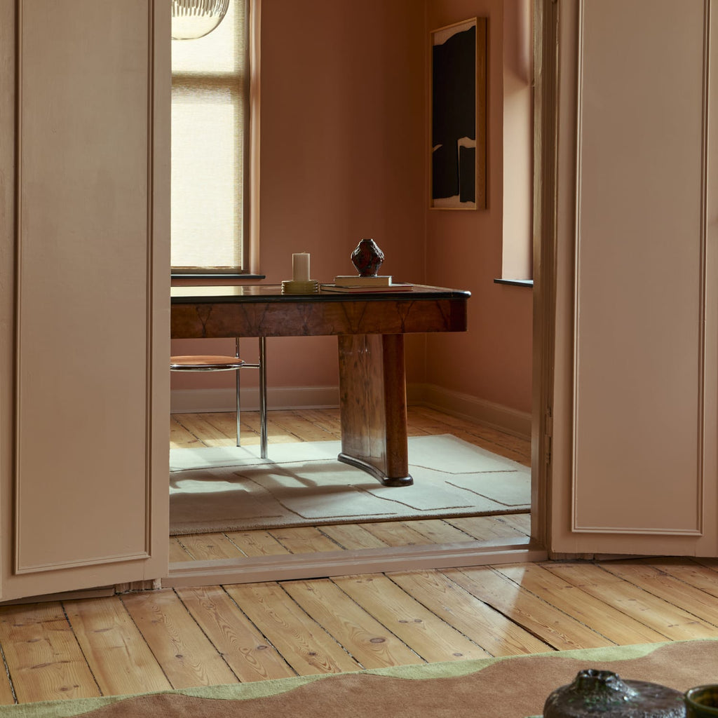 A warm, rustic wooden writing desk in a beautifully styled room with dusty rose walls, a window with a beige roller shade, and a light-colored rug.