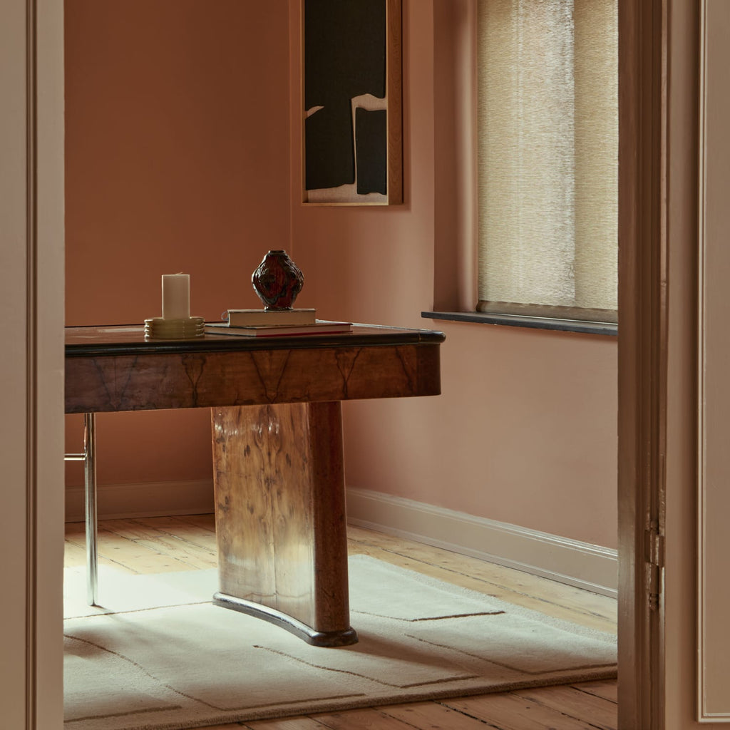 A warm, rustic wooden writing desk in a beautifully styled room with dusty rose walls, a window with a beige roller shade, and a light-colored rug.