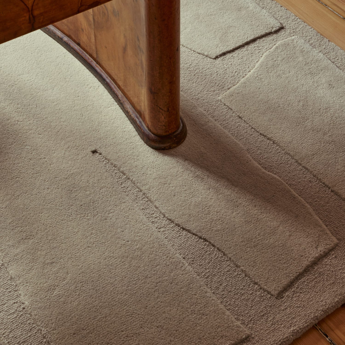 Close-up of a beige area rug with a raised geometric pattern, shown under a wooden table leg on a hardwood floor.