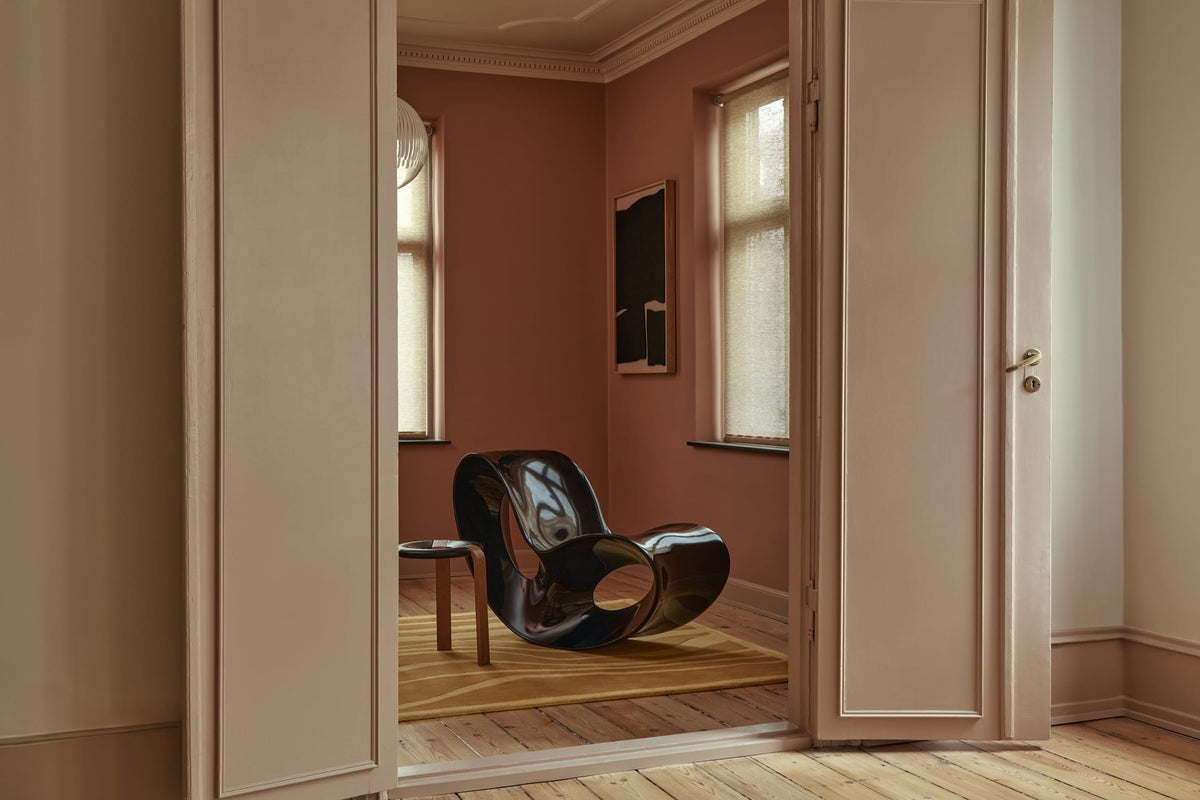 A glossy black sculptural lounge chair sits in a warmly lit room with terracotta walls, wood floors, and open cream-colored doors framing the scene.