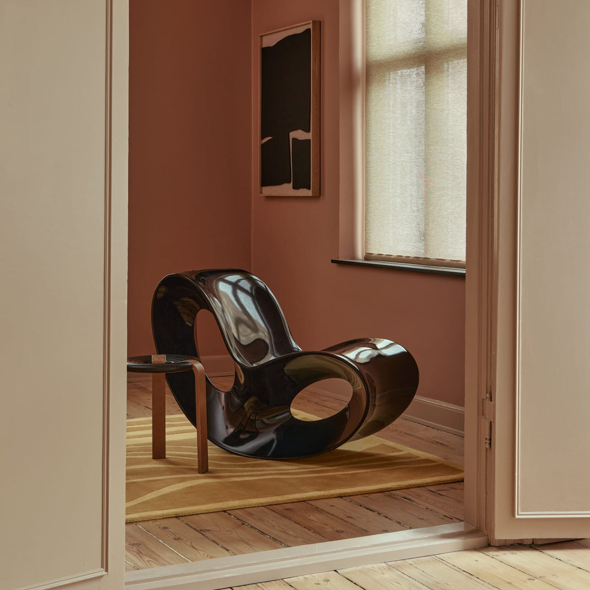 A glossy black sculptural lounge chair with organic looped curves, styled in a warm, minimalist room with terracotta walls, a wooden floor, and a small side table beside it.