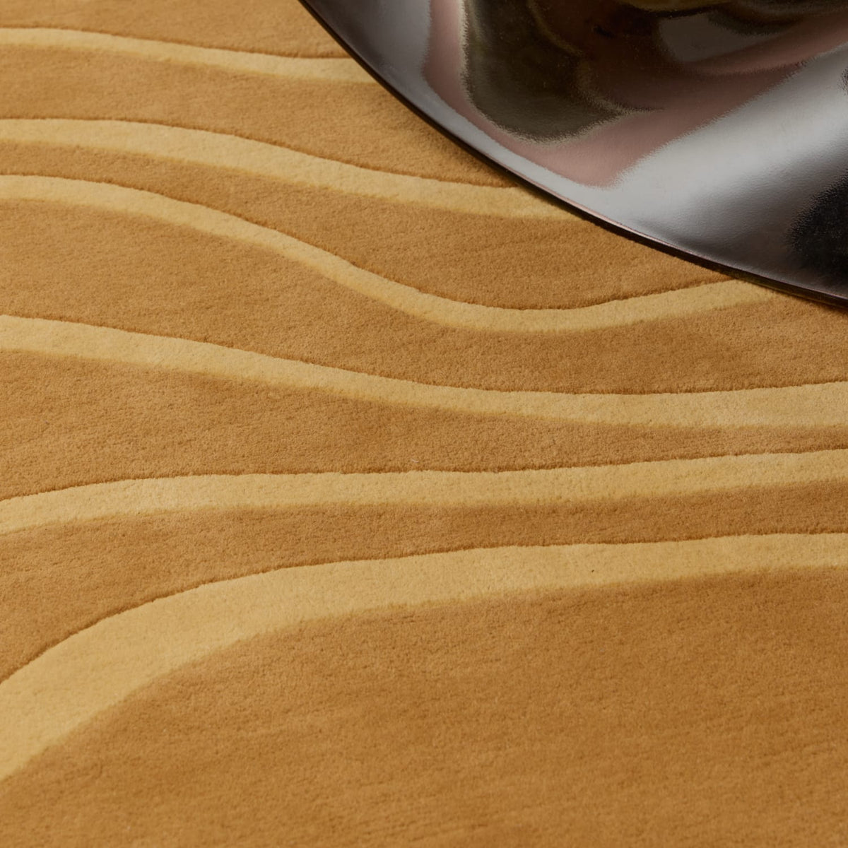 Close-up of a textured tan and beige wavy-patterned surface with a reflective black metallic object partially visible in the top right corner.