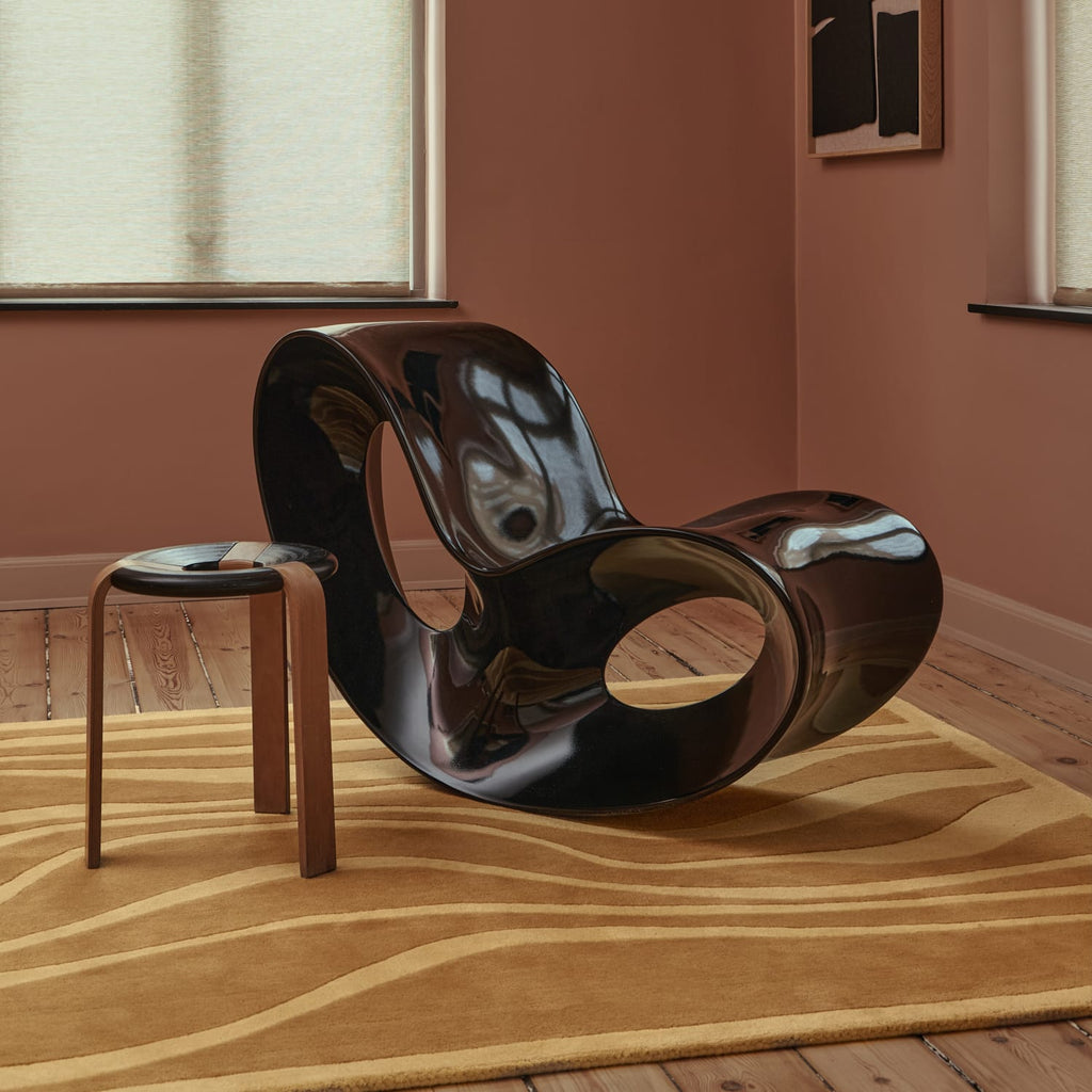 A glossy black sculptural lounge chair with a circular cutout, styled in a room with terracotta walls, a beige patterned rug, and a small wooden stool beside it.