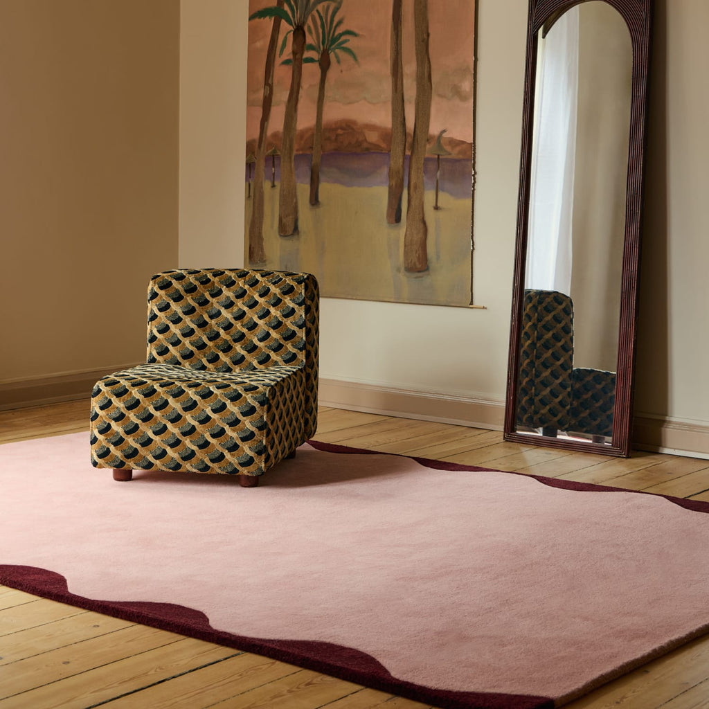 A pink area rug with a dark burgundy wavy border is shown on a light wood floor in a styled room, featuring a patterned armchair and a tall floor mirror.