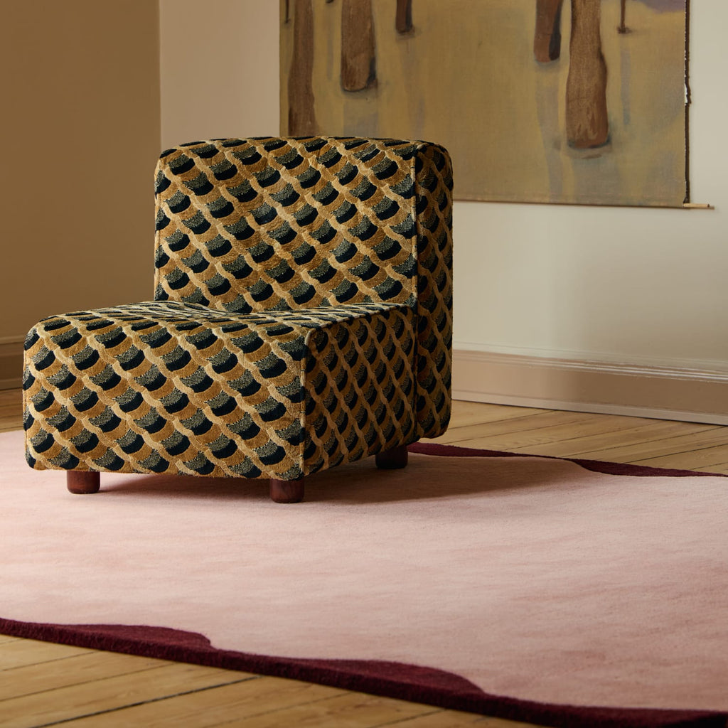 A small upholstered accent chair with a geometric fish-scale pattern in black and tan, placed on a pink area rug in a living room setting.