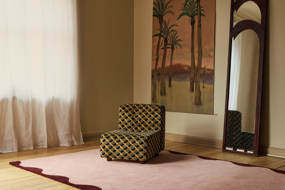 A small upholstered armchair with a black and gold geometric pattern sits on a dusty pink area rug in a styled room. The room features a warm beige wall, a large vertical painting of palm trees, a tall dark wood floor mirror, and sheer white curtains.