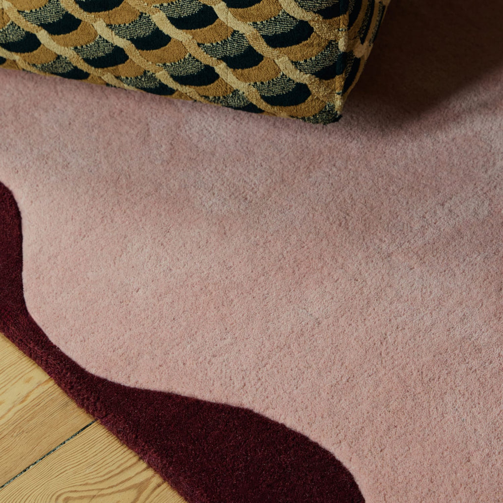 A close-up of a plush, wavy-edged area rug in dusty pink, with a contrasting dark burgundy section and a gold and navy patterned cushion resting on top.