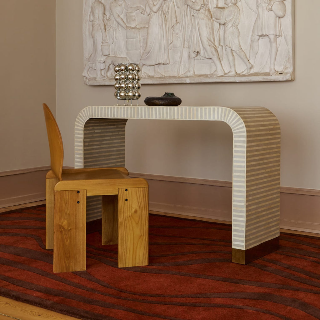 A modern console table with a curved backrest and striped cream-and-brown legs, paired with a light wood stool, set in a room with a red patterned rug and a large white wall sculpture.