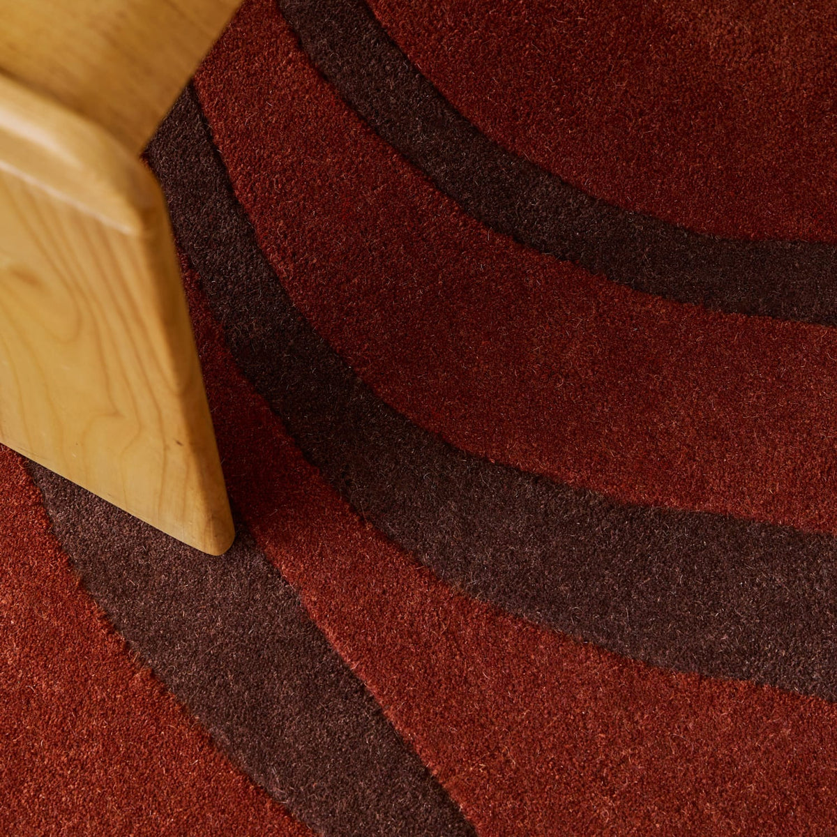 A close-up of a textured area rug with wavy concentric stripes in rust red and dark brown, with a light wood chair leg visible in the upper left corner.